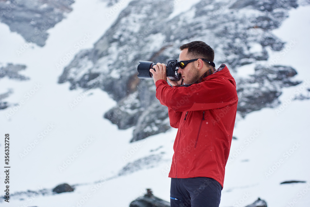 Side view of male photographer taking photo with professional camera. Guy wearing red jacket while photographing winter nature. Concept of tourism and professional photography.