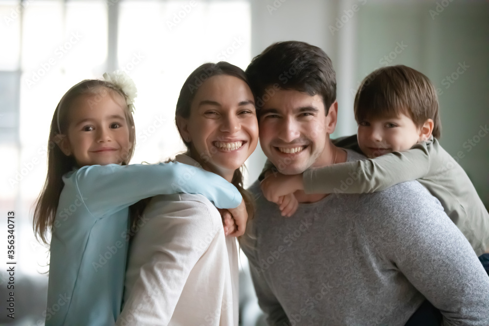 Portrait of smiling Caucasian family with small children relax at home on weekend together, happy little preschooler kids piggyback young parents, play engaged in funny game indoors, look at camera Portrait of smiling Caucasian family with small children relax at home on weekend together, happy little preschooler kids piggyback young parents, play engaged in funny game indoors, look at camera