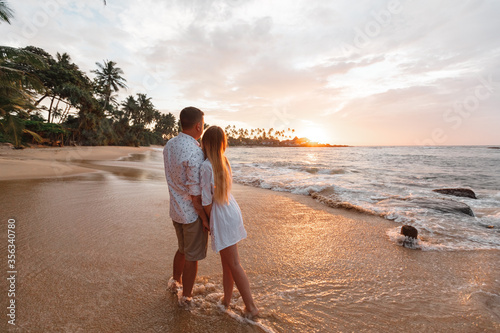 Young happy couple on seashore, looking beautiful golden sunset during vacation on tropical islands, palm trees on background, waves touch their feets