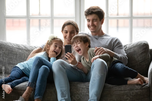 Overjoyed young family with small children sit relax on sofa at home laugh watching funny video on smartphone together, happy Caucasian parents rest with little kids using modern cellphone gadget