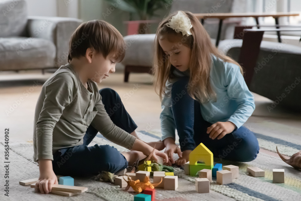 Fototapeta premium Concentrated little brother and sister sit on warm floor in living room play with building bricks or blocks, small preschooler boy and girl children have fun engaged in game with wooden toys at home
