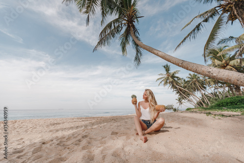 Beautiful woman with long blond hair is holding pineapple fruit on the beach, sitting on the background of ocean and coconut palm trees. Vacation on tropical island