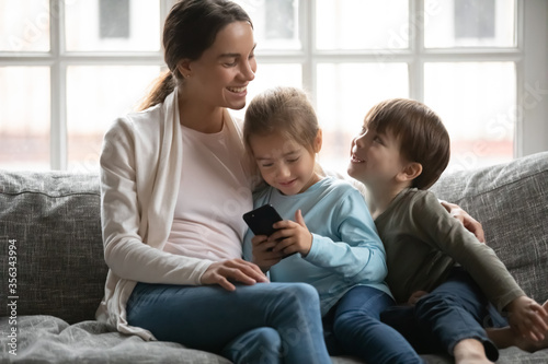 Smiling young Caucasian mom sit relax on sofa with little preschooler children using smartphone together, loving mother rest on couch with small kids, browse internet on modern cellphone gadget