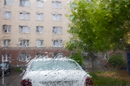 Fuzzy view of the rainy yard through the wet windshield of a car. Tree, automobile and a building wall.