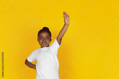 Pulling her arm up, asking a question. Little african-american girl's portrait on yellow studio background. Cheerful kid. Concept of human emotions, expression, sales, ad. Copyspace. Looks cute.