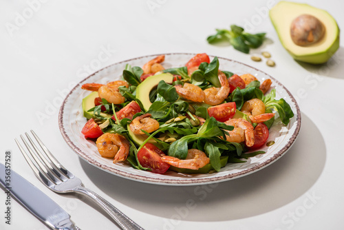 selective focus of fresh green salad with shrimps and avocado on plate near cutlery on white background