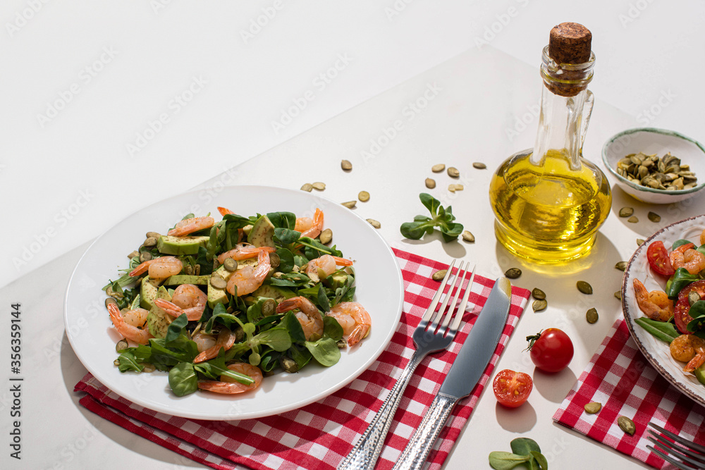 fresh green salad with shrimps and avocado on plate near cutlery on plaid napkin and ingredients on white table