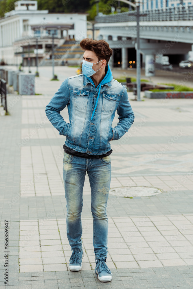 Young man in medical mask and hands in pockets of denim jacket walking on urban street