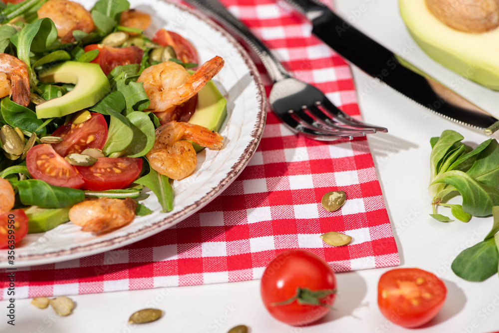 close up view of fresh green salad with shrimps and avocado on plate near cutlery on plaid napkin and ingredients on white background close up view of fresh green salad with shrimps and avocado on plate near cutlery on plaid napkin and ingredients on white background