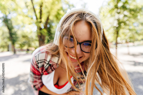 Close-up shot of enthusiastic long-haired girl in headphones. Young lady in glasses fooling around in summer park.