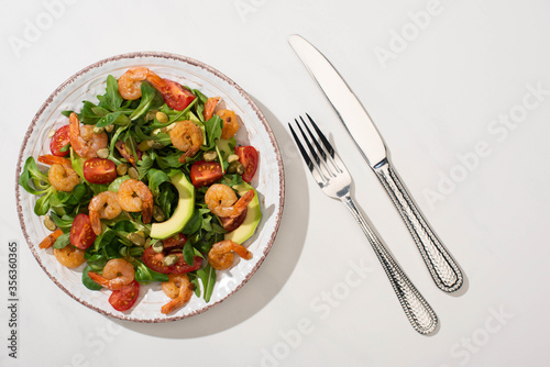top view of fresh green salad with shrimps and avocado on plate near cutlery on white background