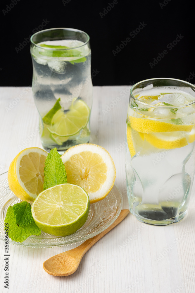 Top view of two glasses of water with lemon and lime slices with ice cubes on white wood, with wooden spoon, plate with half lemons and mint, on black background, vertical Top view of two glasses of water with lemon and lime slices with ice cubes on white wood, with wooden spoon, plate with half lemons and mint, on black background, vertical