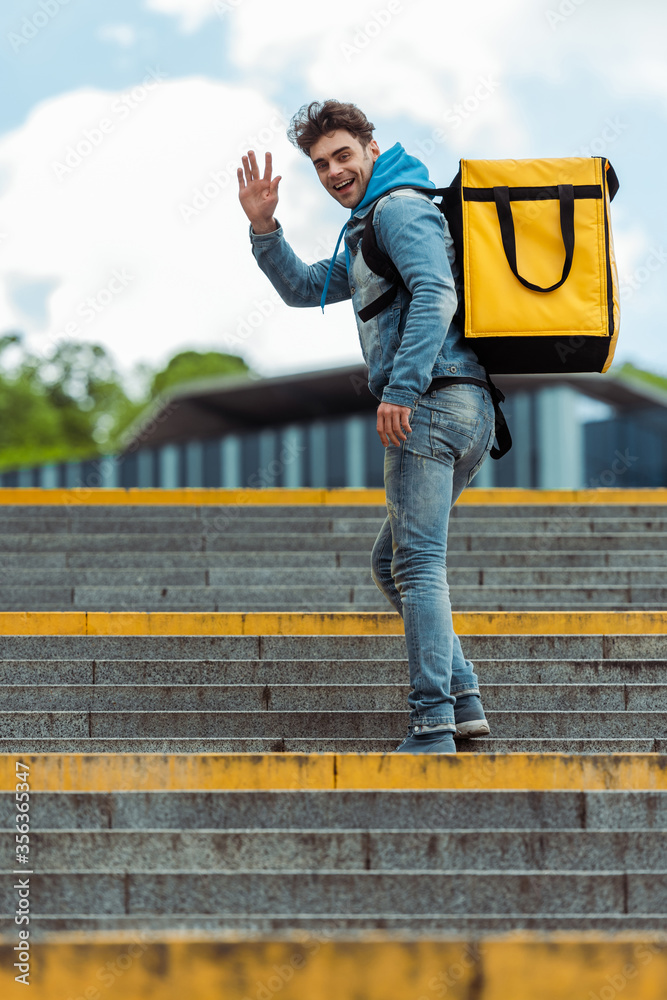Side view of positive delivery man with thermal backpack waving hand at camera on stairs