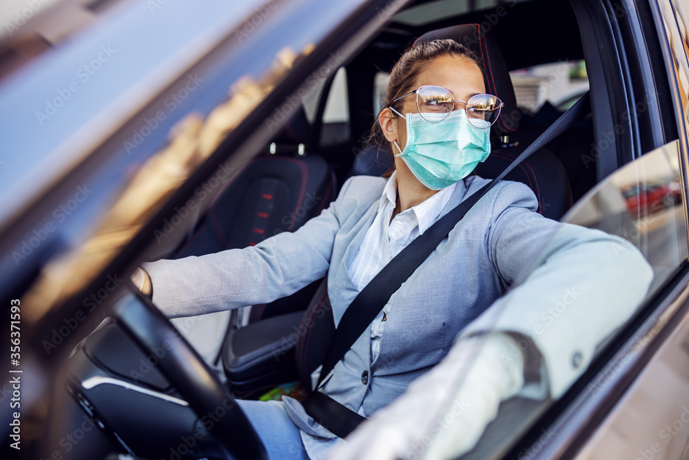 Young attractive businesswoman dressed smart casual with protective mask and gloves on sitting in her car and she is ready to drive it. Protection from corona virus concept.