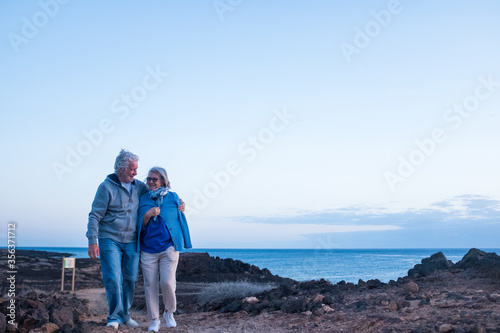 couple of senior walking together isolated on the rocky beach with sea or ocean at the background in the evening - love and affection concept and lifestyle - mature man and woman walking