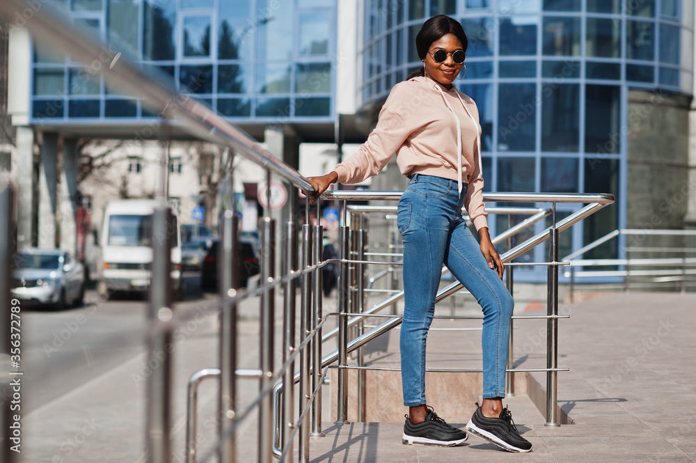 Hipster african american girl wearing pink hoodie,sunglasses and jeans posing at street against office building with blue windows.