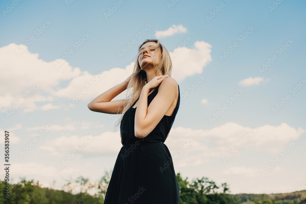 Young blonde attractive woman with long hair, wearing black maxi dress, outdoors, standing in a field, on blue sky background. Young blonde attractive woman with long hair, wearing black maxi dress, outdoors, standing in a field, on blue sky background.