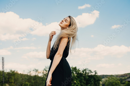 Young blonde attractive woman with long hair, wearing black maxi dress, outdoors, standing in a field in wind, on blue sky background.