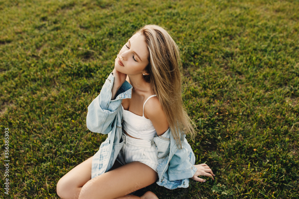 Young attractive woman with long blonde hair, with eyes closed, wearing a denim jacket, sitting on green grass. Young attractive woman with long blonde hair, with eyes closed, wearing a denim jacket, sitting on green grass.