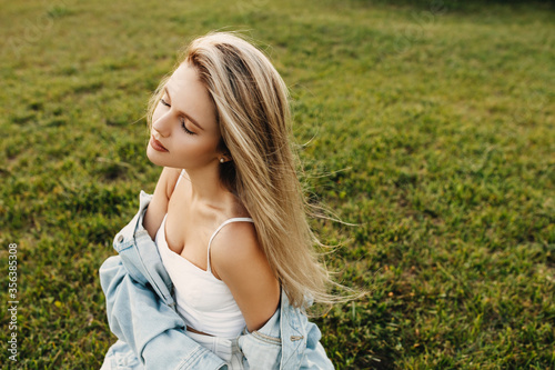 Young attractive woman with long blonde hair, with wind in hair, wearing a denim jacket, sitting on green grass.