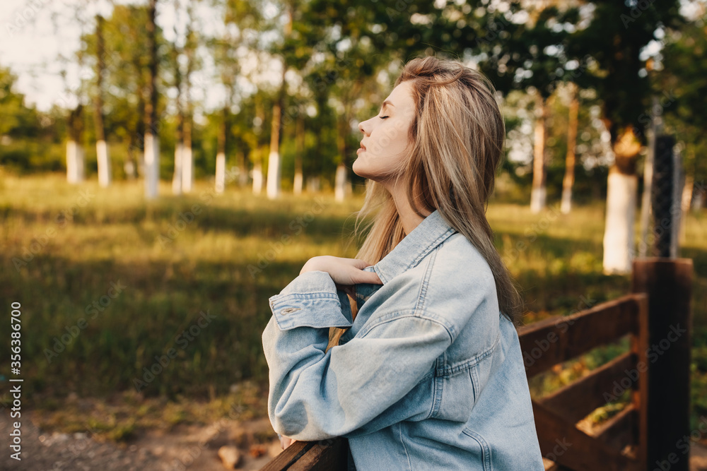 Young woman with long blonde hair, in golden hour, outdoors, wearing a denim jacket, standing by a wooden fence.