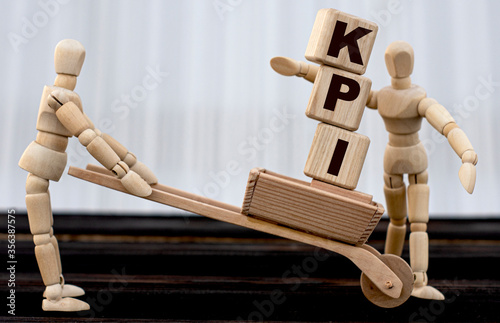 KPI - acronym on cubes standing in wheelbarrow. A wooden man passes them to the second person. photo