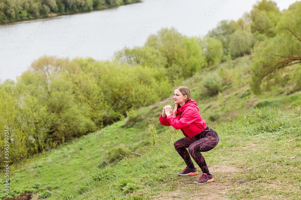 Athletic caucasian woman doing squats on the river bank outdoors. Sporty lifestyle. Beautiful girl is doing fitness exercises outside in the park. Athletic caucasian woman doing squats on the river bank outdoors. Sporty lifestyle. Beautiful girl is doing fitness exercises outside in the park.