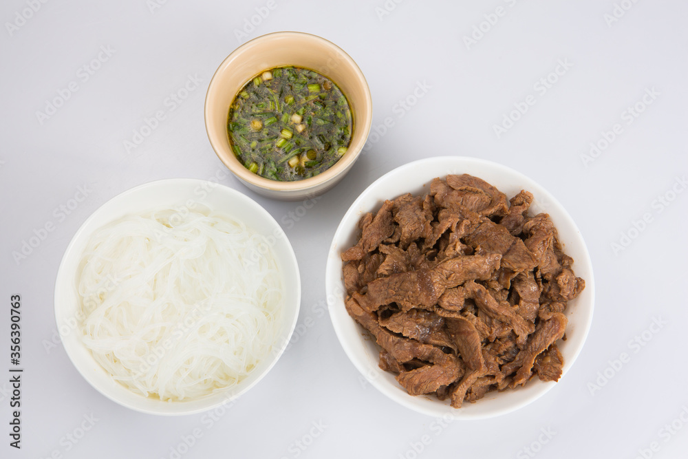 isolated close up flat lay top view shot of a bowl of sliced beef fillet pieces, a plate of cellophane glass noodles and a cup of Vietnamese Nuoc Cham dipping sauce on a white background isolated close up flat lay top view shot of a bowl of sliced beef fillet pieces, a plate of cellophane glass noodles and a cup of Vietnamese Nuoc Cham dipping sauce on a white background