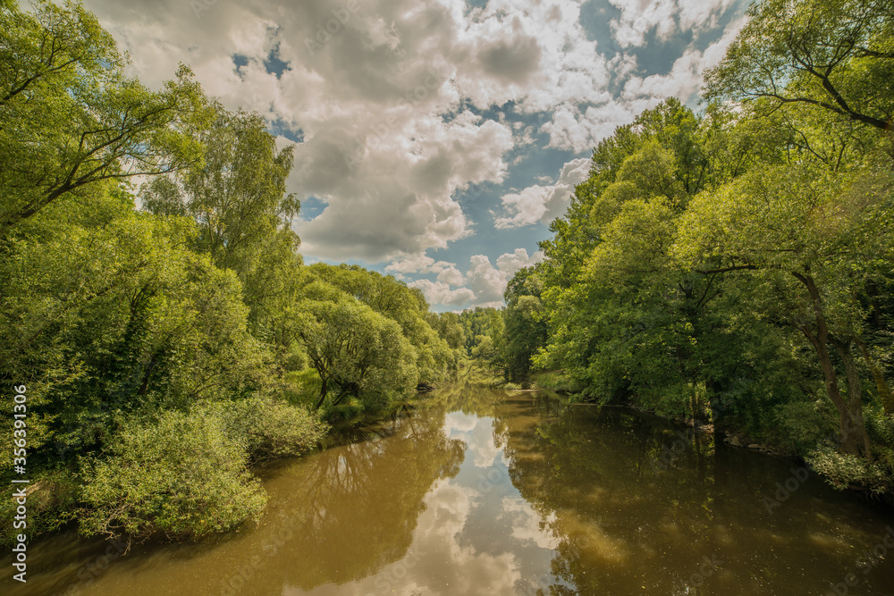 view from a bridge on calm river surrounded by green trees and mirroring blue sky with clouds