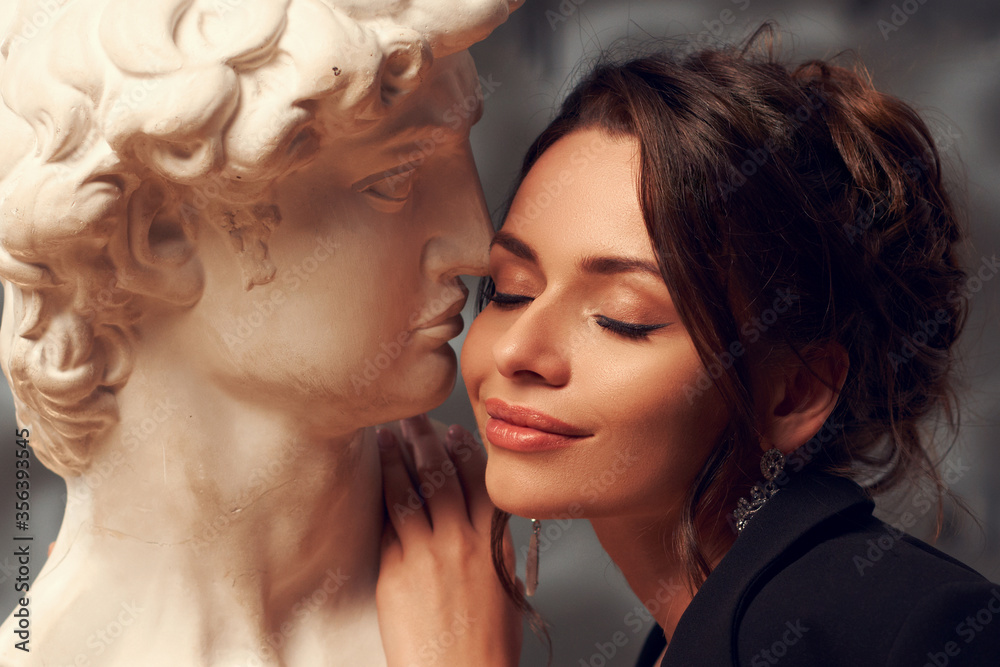 Fashion style portrait of young elegant beautiful caucasian woman with hairstyle and makeup kissing with gypsum head. Love concept