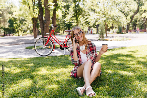 Jocund woman in summer clothes sitting on grass and drinking coffee. Outdoor shot of fascinating girl in glasses talking on phone on nature background.