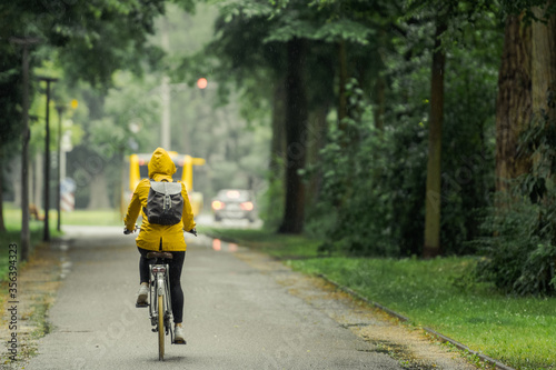 One unrecognizable person with yellow oilskin on bike riding through rainy street in beautiful old alley with trees on rainy day