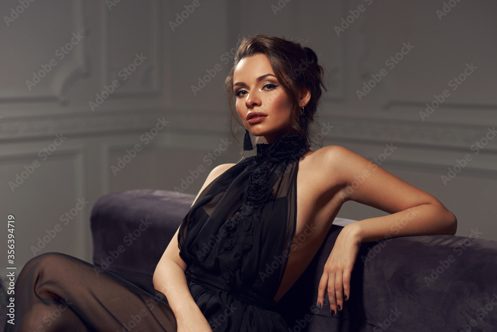 Pretty stylish girl in black dress with makeup and hairstyle sitting at gray sofa in luxury interior. Fashion style portrait Pretty stylish girl in black dress with makeup and hairstyle sitting at gray sofa in luxury interior. Fashion style portrait