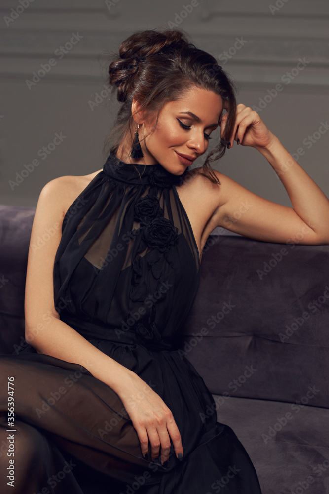 Pretty stylish girl in black dress with makeup and hairstyle sitting at gray sofa in luxury interior. Fashion style portrait Pretty stylish girl in black dress with makeup and hairstyle sitting at gray sofa in luxury interior. Fashion style portrait