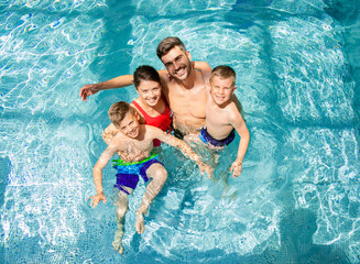 Top view of smiling family of four having fun and relaxing in indoor swimming pool at hotel resort.
