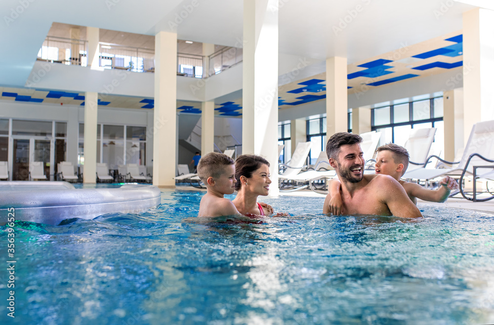 Smiling family of four having fun and relaxing in indoor swimming pool at hotel resort.