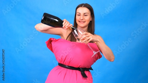 The most beautiful girl in a pink dress from pillows pours champagne into a glass on a blue background. Crazy quarantine. Fashion 2020. Put on a pillow. Challenge 2020 due to house isolation. photo