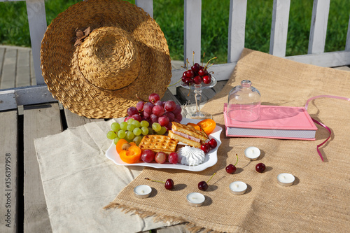 Still life and food photo. Picnic in nature on a sunny day. A straw hat and a dish of fruit, berry and waffles stand on a burlap fabric on a wooden floor from old boards