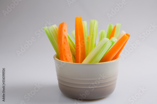 close up filled frame isolated side view shot of a brown beige bowl filled with crunchy orange carrot slices and juicy green celery sticks on a white background