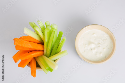 close up isolated flat lay top view shot of party snack food. A bowl of crunchy orange carrot and juicy green celery sticks with a white cup of blue cheese dipping sauce on a white background