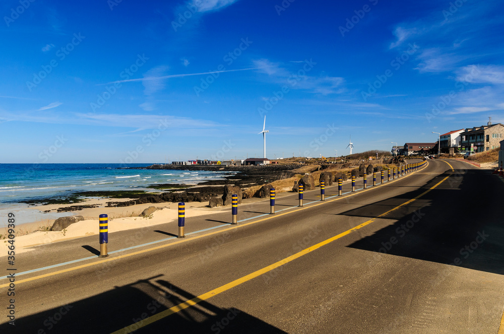 Beautiful view Woljeong Beach besides trunk road providing leading line at Jeju Island South Korea with white windmill at far left backed by awesome blue sky background