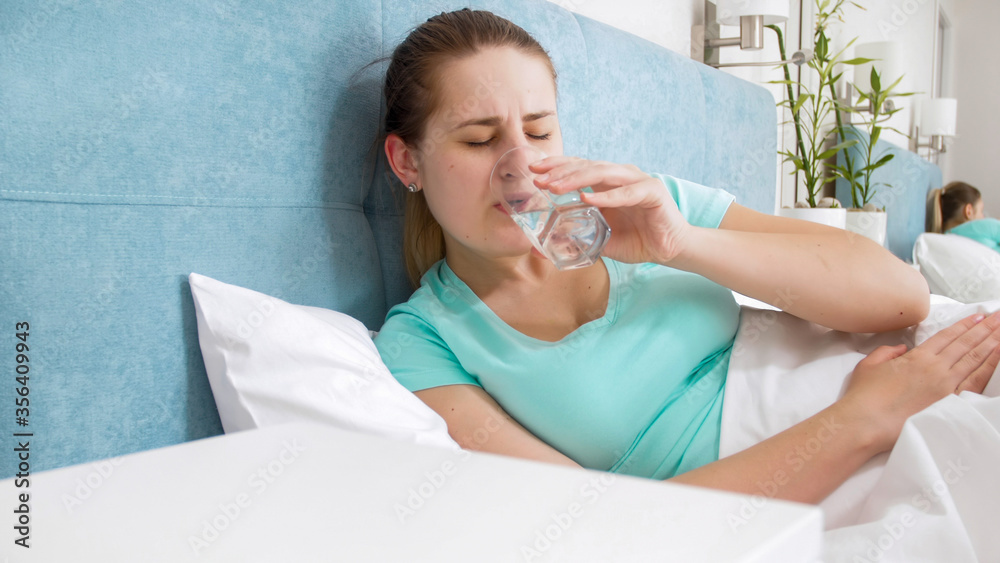 Portrait of young woman feeling unwell lying in bed at home and drinking water Portrait of young woman feeling unwell lying in bed at home and drinking water