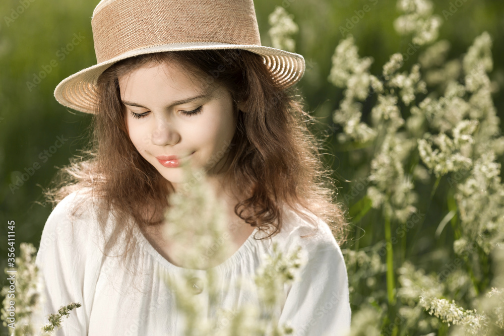 The portrait of a cute girl in a straw hat in summer morning, she is smiling. Cheerful female kid in straw hat and white dress enjoying vacation spending time.