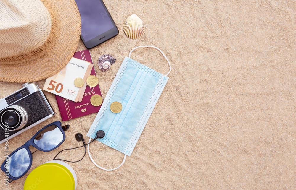 Some beach items, a protective face mask, a passport and some cash, a smartphone, hat, camera and sunglasses on the beach sand. Space for text. Concept of summer in the covid-19 period. Some beach items, a protective face mask, a passport and some cash, a smartphone, hat, camera and sunglasses on the beach sand. Space for text. Concept of summer in the covid-19 period.