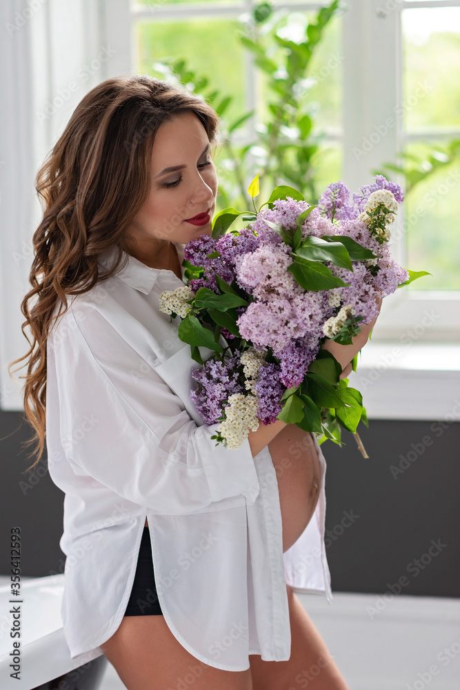 Beautiful pregnant woman in a white shirt in the bathroom near the window with a bouquet of lilac flowers. Soft selective focus. Beautiful pregnant woman in a white shirt in the bathroom near the window with a bouquet of lilac flowers. Soft selective focus.