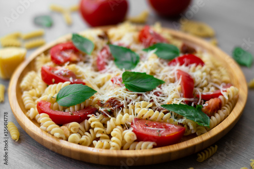 Summer cold pasta salad with fusilli pasta, fresh tomatoes, sun-dried tomatoes, grated Parmesan cheese and fresh green basil leaves in a wooden plate surrounded by tomatoes, basil, dry pasta and parm