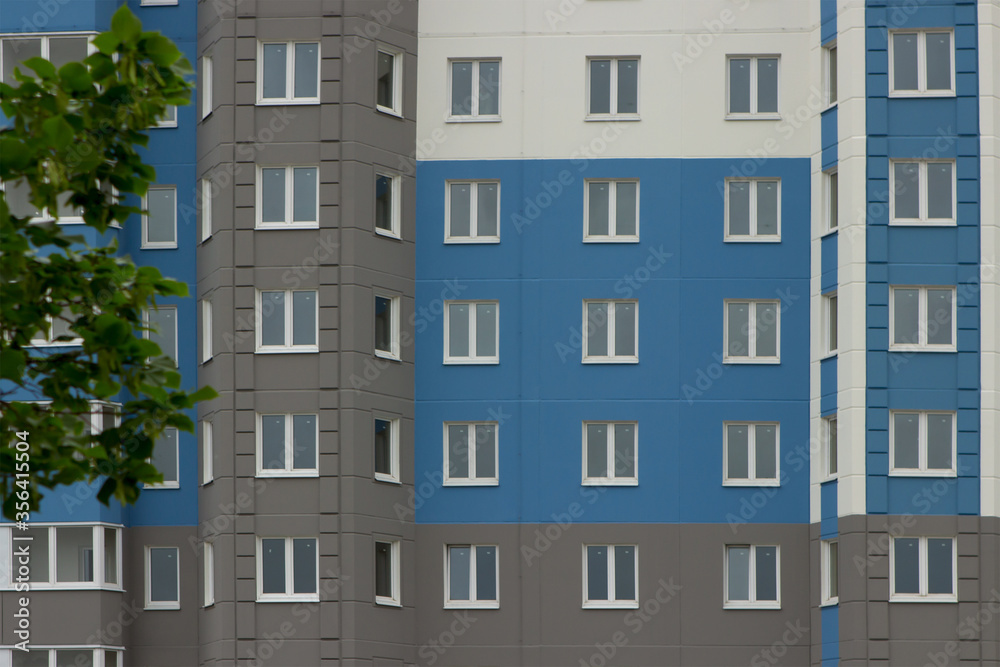 A modern, newly-built multi-apartment multi-storey residential building in which no one lives and apartments are for sale. The facade of a blue building with still uninhabited windows against a backgr A modern, newly-built multi-apartment multi-storey residential building in which no one lives and apartments are for sale. The facade of a blue building with still uninhabited windows against a backgr