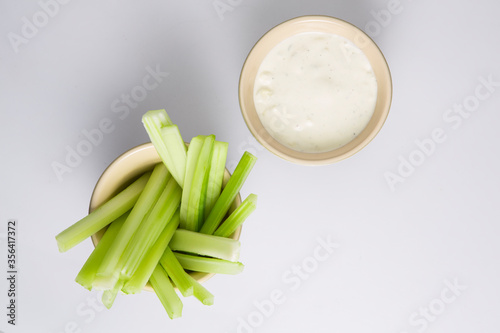 close up isolated flat lay top view shot of a bowl of crunchy juicy green celery sticks next to a white cup of blue cheese dipping sauce on a white background