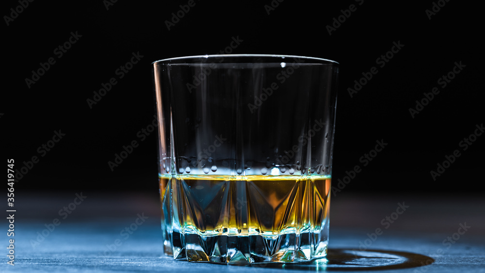 A glass of whiskey lit by a ray of light stands on a dark table and on a black background