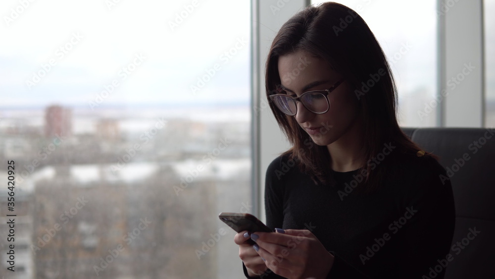 A young woman with a phone. A girl sits in a cafe by the panoramic window with a smartphone in her hands. View from the window. A young woman with a phone. A girl sits in a cafe by the panoramic window with a smartphone in her hands. View from the window.